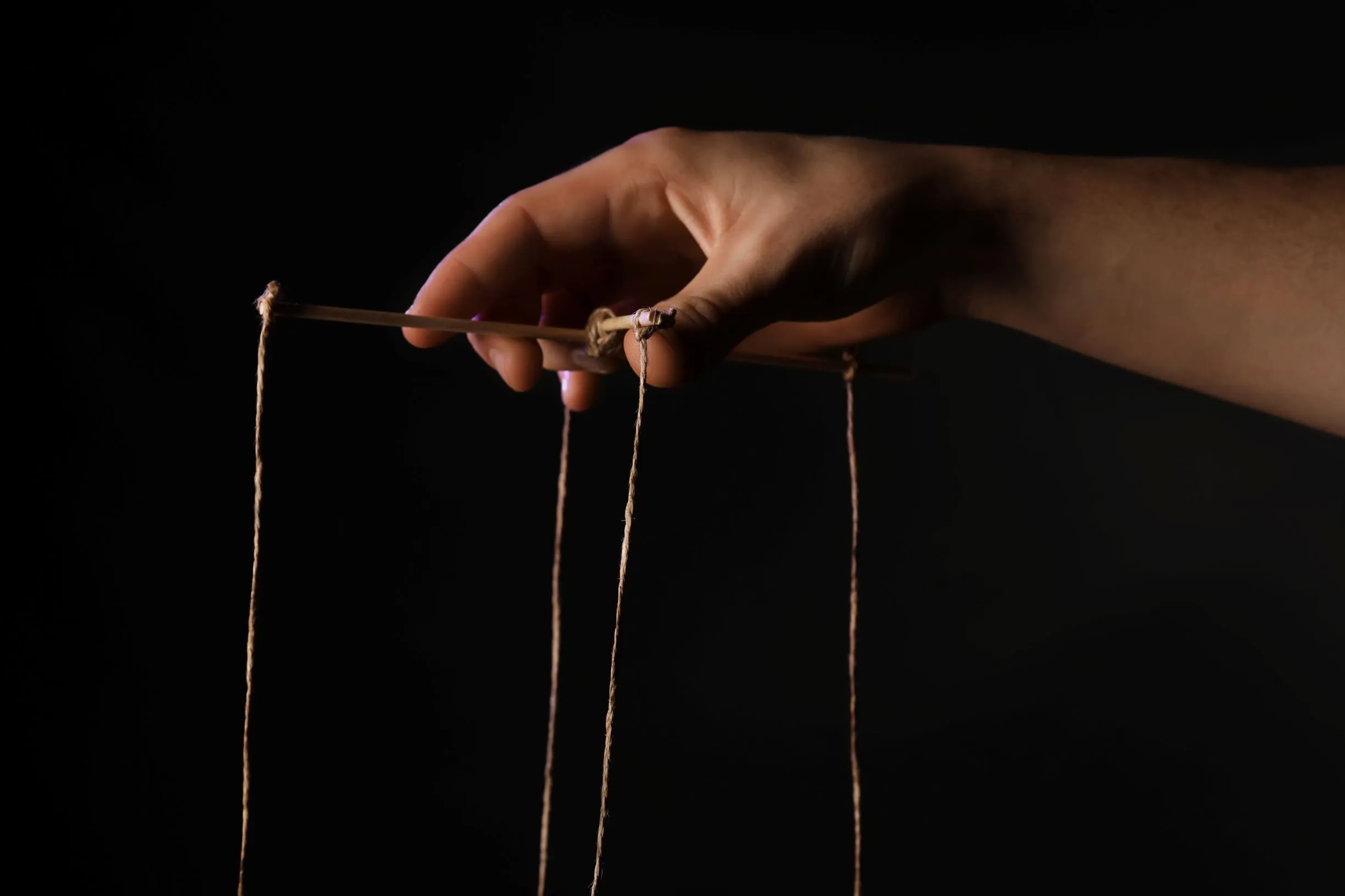 A hand working the strings of a marionette against a black background